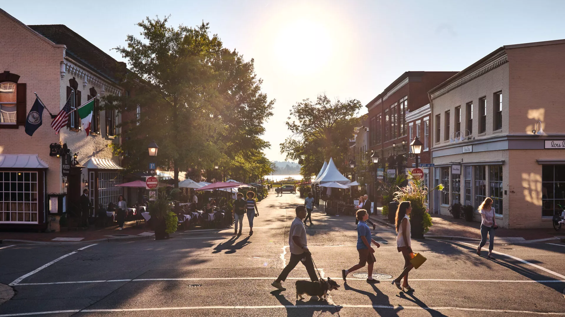 People cross a sunlit street in a charming downtown area lined with shops, restaurants, and outdoor seating. Trees provide shade and the sun is low in the sky, creating a warm and inviting atmosphere.