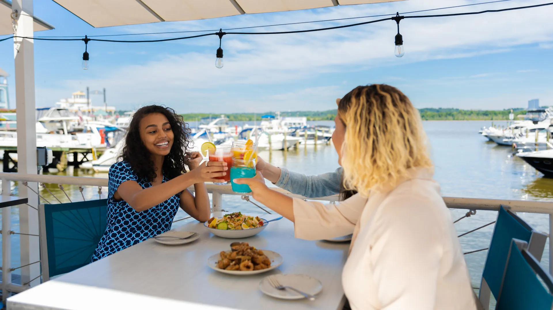 Three people sit at an outdoor table by a marina, smiling and toasting colorful drinks. Plates of food are on the table, and boats are docked in the background under a sunny, blue sky.
