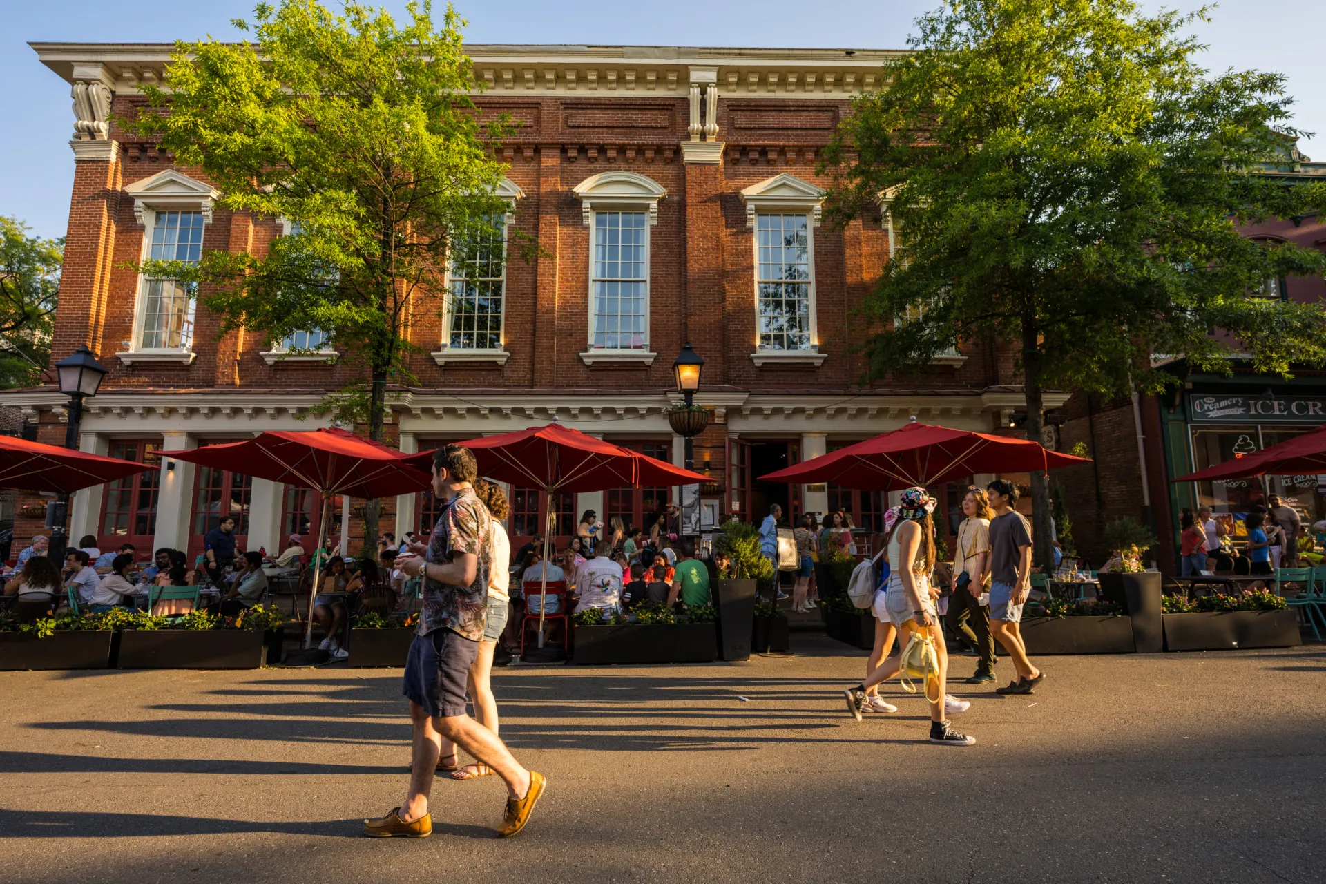 People walk past a busy outdoor restaurant with red umbrellas on a sunny day. The scene is set in front of a historic brick building with large windows and trees lining the sidewalk.