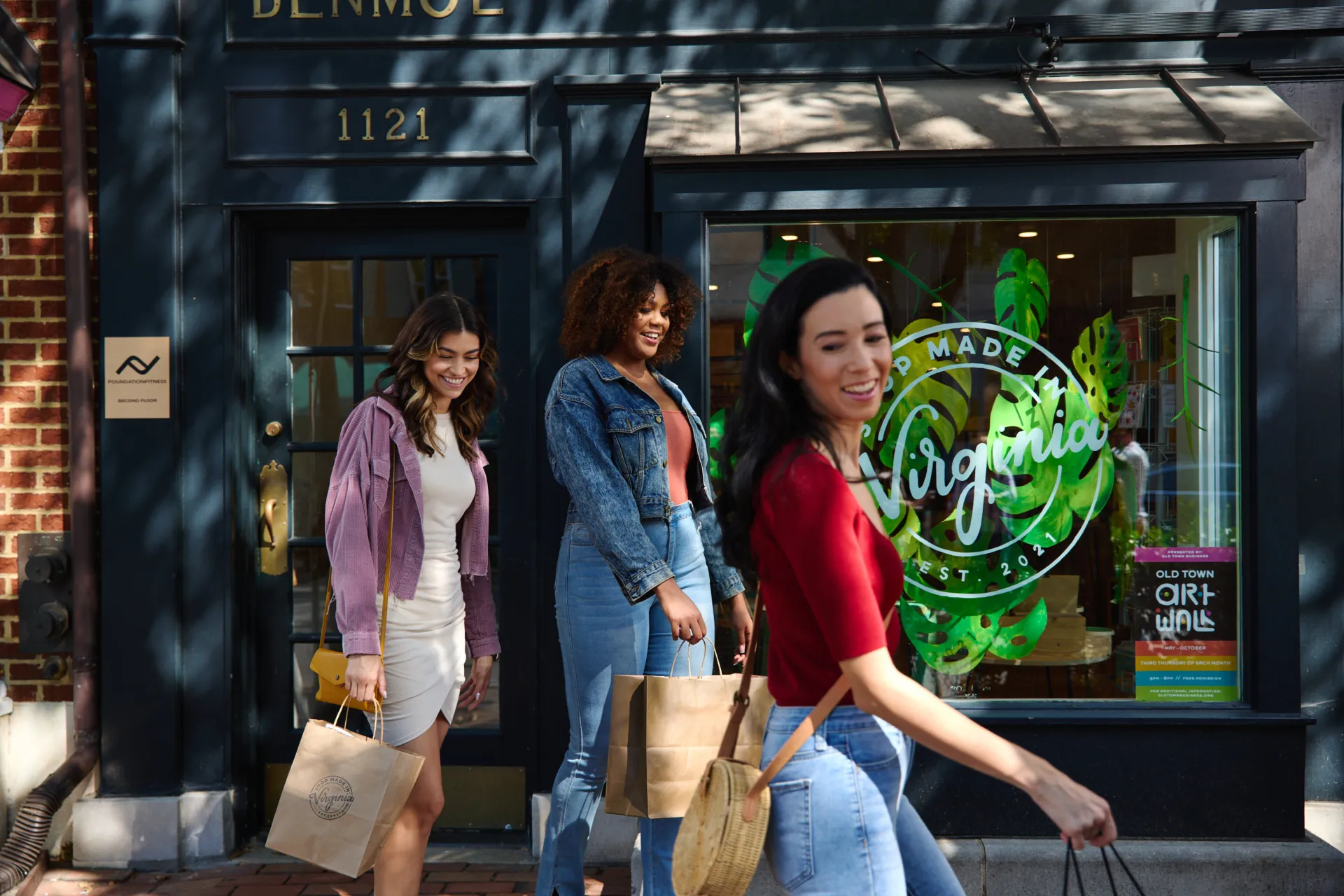 Three women smiling and carrying shopping bags walk past a storefront with Made in Virginia on the window. Brick buildings and sunlit trees are visible along the street.