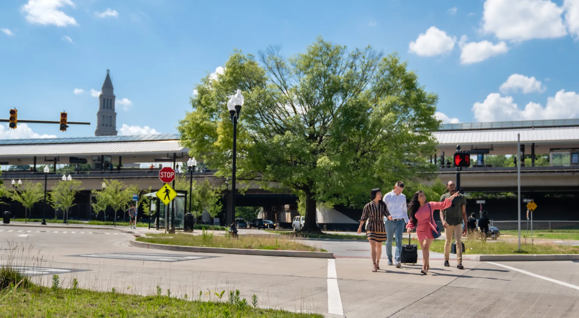 A group of five people, including two children, cross a street at a crosswalk on a sunny day. A large tree, a train station, and a clock tower are visible in the background under a blue sky with scattered clouds.