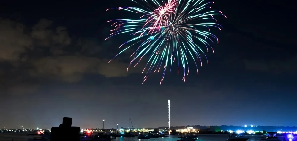 Colorful fireworks light up the night sky over a body of water, with boats and city lights visible on the horizon below.